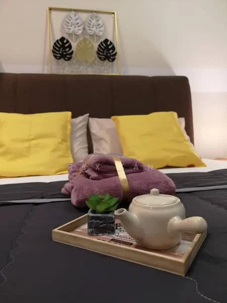 Bedroom features: queen-size bed with brown upholstered headboard, yellow square throw pillows, grey bedspread, decorative wall art with leaves, small potted plant on tray, white ceramic teapot, wooden tray, purple towel, soft lighting, neutral wall color; style: modern.