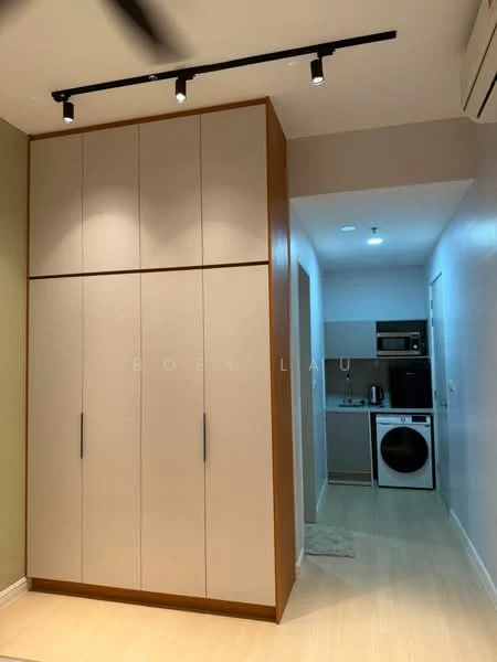 Room features: built-in wardrobe with light wood and beige finish, track lighting with multiple spotlights, light-colored laminate flooring, open kitchen area visible in the background, kitchen appliances including fridge and washing machine, neutral wall color, air conditioning unit on the wall; style: modern.