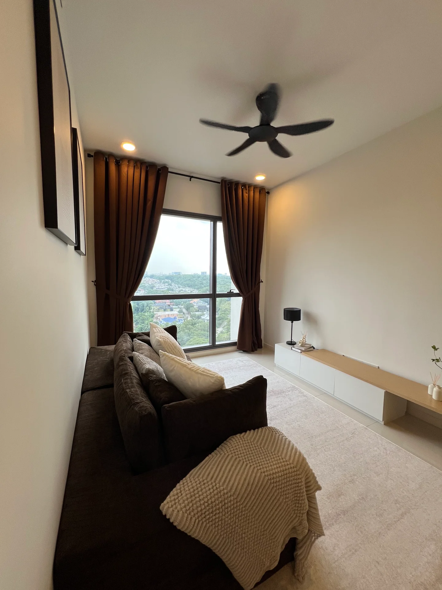Living features: 3-seater fabric sofa in brown, 2 decorative cushions, floor-to-ceiling windows, sheer curtains, blackout curtains, ceiling fan with light, light-colored wall, carpet flooring, TV console, decorative plant on console; style: modern; view: city.