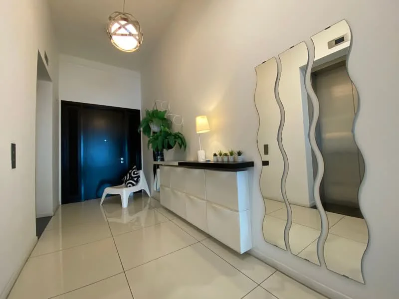Hallway features: white painted walls, glossy tile flooring, modern pendant light, elevator access, large mirror with wavy design, console table with black top and white drawers, decorative houseplants, white accent chair; style: modern.