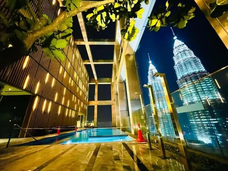 Exterior type: high-rise condominium; features: infinity pool, pool deck with loungers, glass railing, decorative tree, night lighting, city skyline view, Petronas Twin Towers in background; style: modern.