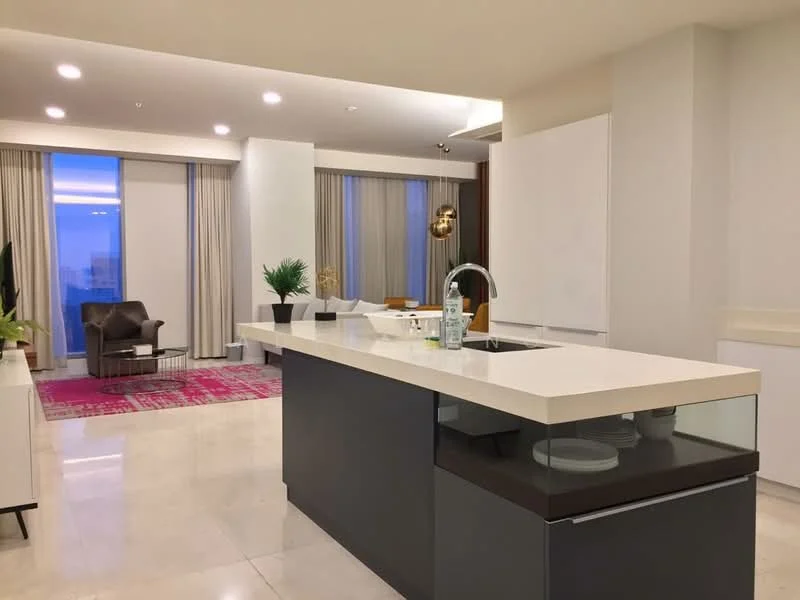 Kitchen features: island kitchen with white countertop, stainless-steel sink, chrome pull-down faucet, built-in cabinetry, white cabinetry, marble flooring, pendant light fixture, floor-to-ceiling windows, curtains, decorative plant, area rug with geometric pattern, seating area with armchair; style: modern; view: city.