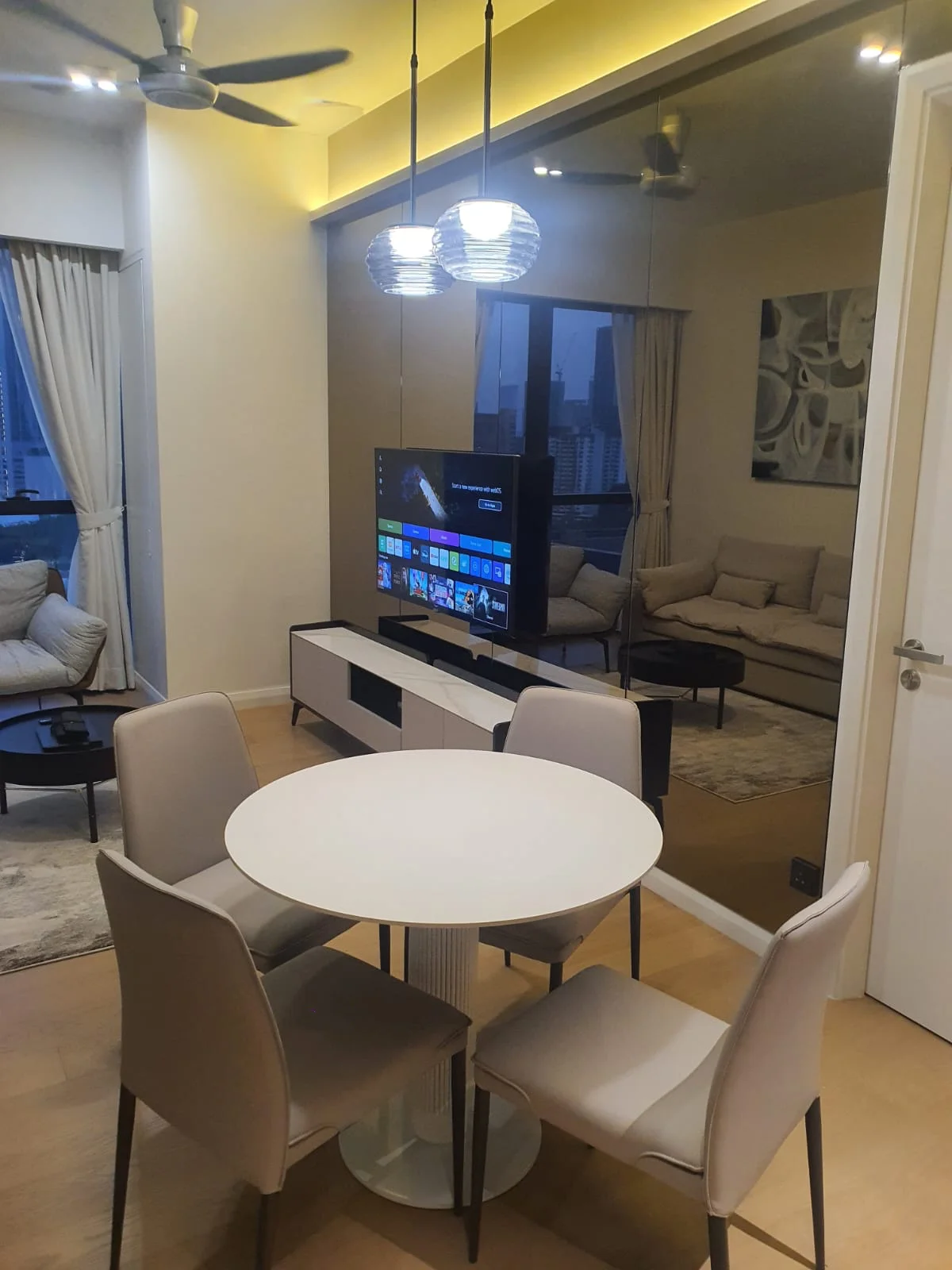 Dining features: round dining table, 4 upholstered dining chairs, pendant light fixture, wall-mounted TV, light wood flooring, floor-to-ceiling windows, sheer curtains, mirrored wall, sofa in background, area rug; style: modern; view: city.