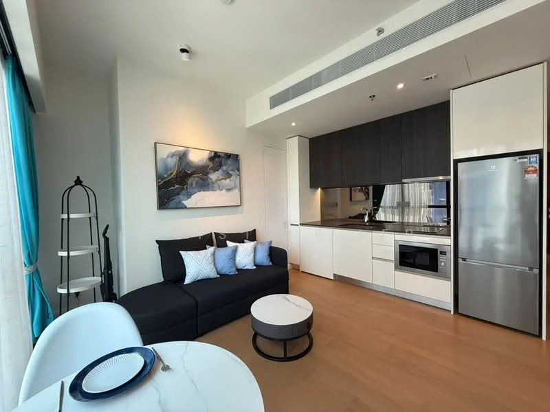 Living features: L-shape sectional sofa in black, decorative cushions in various shades of blue and white, round coffee table with white marble top, built-in kitchen cabinetry in white, dark wood upper cabinets, stainless-steel fridge, built-in microwave, ceramic cooktop, kitchen countertop in dark stone, pendant lighting, floor-to-ceiling windows with sheer curtains, light wood flooring; style: modern; view: city.