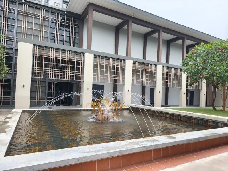 Exterior type: low-rise apartment block; features: water fountain with multiple jets, manicured lawn, landscaped garden with trees, modern architectural design, glass windows, wooden accents, outdoor seating area, paved walkway; style: modern.