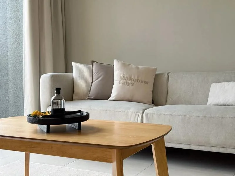 Living features: light grey fabric sofa, decorative throw pillows with text, wooden coffee table, glass bottle on coffee table, small black serving tray on coffee table, neutral painted walls, floor-to-ceiling window with sheer curtains, light-colored tile flooring; style: modern.