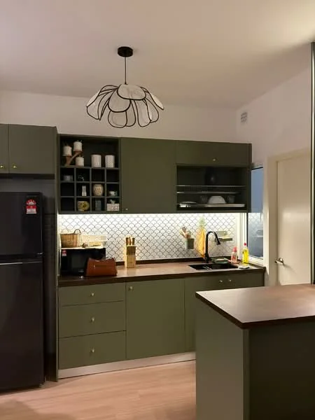 Kitchen features: green cabinetry, white backsplash with hexagonal tiles, black countertop, stainless-steel sink, chrome pull-down faucet, built-in oven, refrigerator, pendant light fixture, open shelving, decorative items on shelves, wooden cutting board, kitchen utensils, drawer handles, light wood flooring; style: modern.