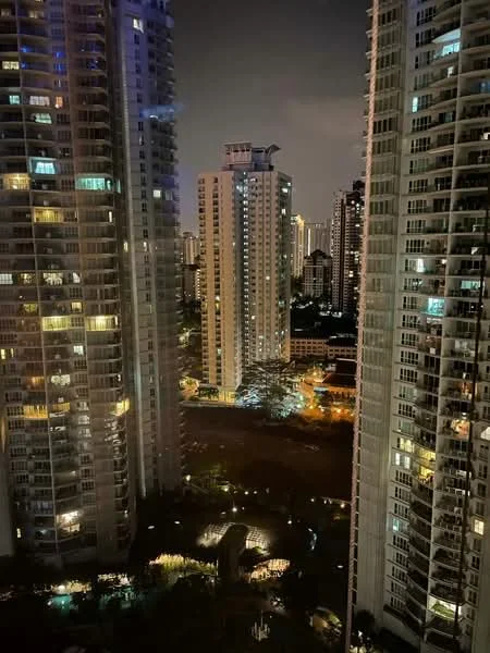 Exterior type: high-rise condominium; features: glass curtain wall, illuminated facade, surrounded by other high-rise buildings, night view, urban skyline, landscaped grounds, parking area visible, city lights, multiple balconies, varied architectural designs; style: modern.