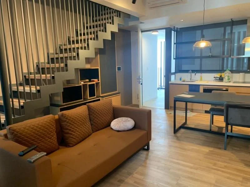 Living features: 3-seater fabric sofa in brown, coffee table, TV console, wooden flooring, staircase with metal railing, pendant lighting, wall-mounted TV, decorative wall paneling; style: modern. Kitchen features: kitchen island, built-in cabinets, countertop, stainless-steel sink, kitchen appliances visible, open shelving; style: modern.