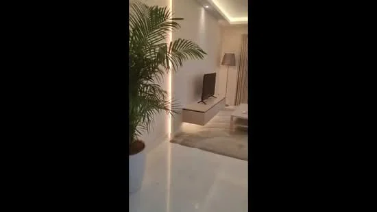 Living features: white painted walls, marble flooring, recessed LED downlights, floor-to-ceiling windows, light grey curtains, TV mounted on wall, light wood TV console, area rug with neutral tones, decorative indoor plant in white pot, table lamp with fabric shade, coffee table with light wood finish; style: modern.