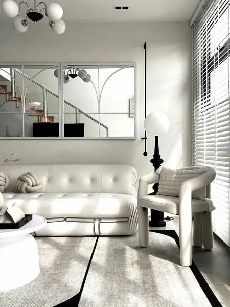 Living features: white tufted sofa, round coffee table, floor lamp with globe shade, decorative throw pillows, area rug with geometric pattern, large mirror with decorative frame, wooden staircase in background, window with blinds, wall-mounted light fixture, decorative plant; style: modern.