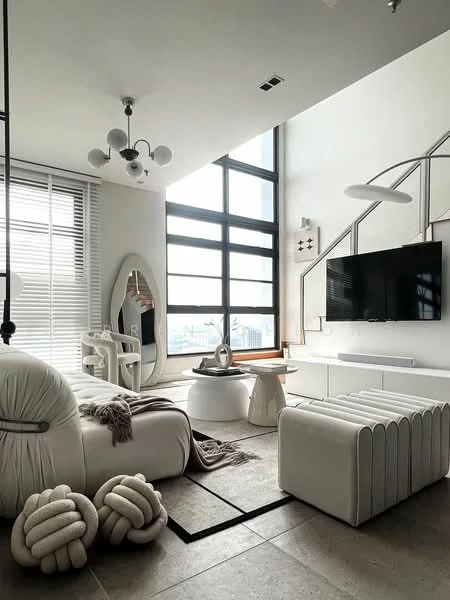 Living features: floor-to-ceiling windows, white walls, grey area rug, 3-seater white sofa, decorative throw blanket, round coffee table, white side table, wall-mounted TV, chandelier with multiple bulbs, large mirror, decorative knot pillows, wooden console table, minimalist decor, natural light from windows; style: modern; view: city.