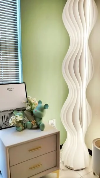 Room features: green painted walls, white floor lamp with wavy design, nightstand with two drawers and gold handles, decorative figurine in green, small vase with flowers, white ceramic pot, window with blinds, tile flooring; style: modern.