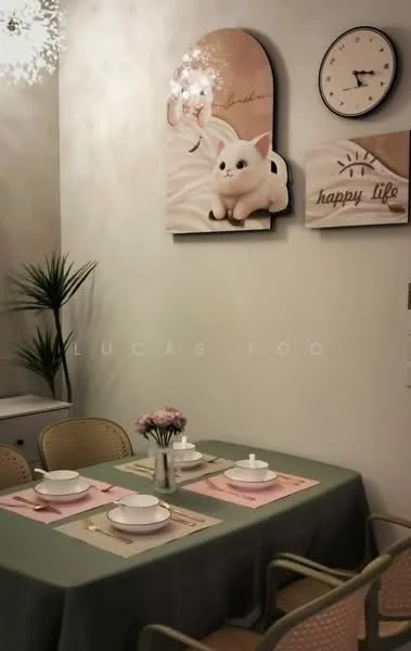 Dining features: 6-seater dining table with green tablecloth, white ceramic plates, pink placemats, decorative flower vase with flowers, wall clock, framed artwork with a pig, framed artwork with the text 'happy life', indoor plant, chandelier lighting, wooden chairs with cane backing; style: modern.
