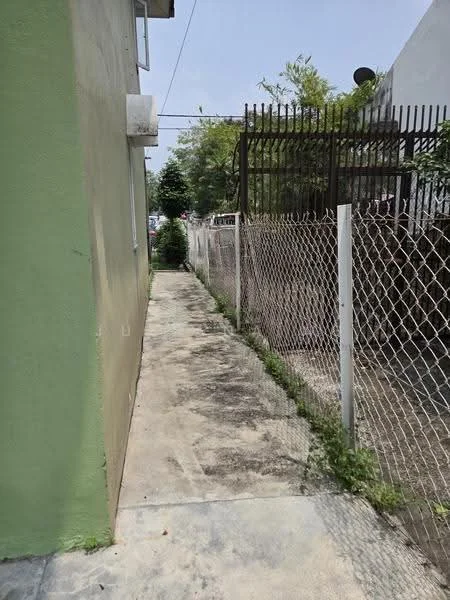 Exterior features: narrow pathway, concrete flooring, green painted wall, fence with chain-link, vegetation along the pathway, adjacent building visible.
