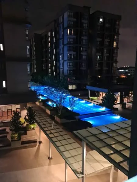 Exterior type: high-rise condominium; features: illuminated infinity pool, landscaped garden with trees, covered walkway with glass roof, outdoor seating area, modern architecture, night lighting, surrounding high-rise buildings; style: modern.