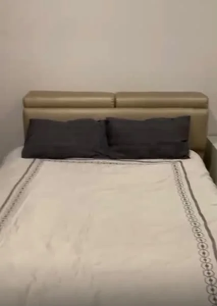 Bedroom features: queen-size bed, upholstered headboard in beige, two grey pillows, white bedspread with decorative pattern, nightstand, plain white walls; style: modern.