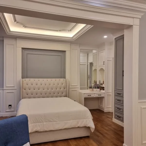 Bedroom features: king-size bed with upholstered headboard, white bedding, built-in wardrobe with grey doors, dressing table with mirror, wooden flooring, tray ceiling with recessed lighting, decorative wall paneling, nightstand with drawers; style: modern.