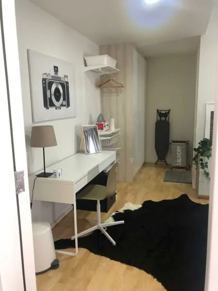 Study features: white desk, ergonomic office chair, black area rug, framed artwork on wall, shelf above desk, hanging clothes rack, ironing board, decorative plant, wooden flooring, white walls; style: modern.