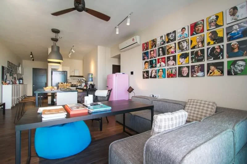 Living features: grey fabric sofa, blue bean bag, black dining table, wooden flooring, ceiling fan, wall-mounted air conditioning unit, artwork on wall, pink fridge, pendant lighting, open layout, decorative wall with framed photos; style: modern.