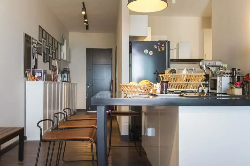 Kitchen features: black countertop, stainless-steel sink, chrome pull-down faucet, built-in oven, freestanding range oven, black refrigerator, wooden cabinetry, open shelving, bar stools, pendant lighting, dark wood flooring, wall-mounted artwork, door leading to hallway; style: modern.