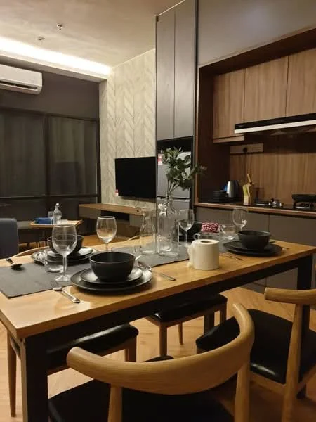 Dining features: wooden dining table, 4 dining chairs, black ceramic plates, glass wine glasses, glass water bottles, cutlery set, table placemats, decorative vase with flowers, wall-mounted TV, kitchen cabinetry, stainless-steel kettle, built-in stove, refrigerator, wooden flooring, recessed lighting; style: modern.