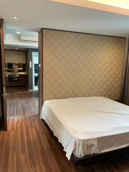 Bedroom features: queen-size bed frame, upholstered headboard with diamond pattern, wooden flooring, recessed ceiling lights, wall-mounted air conditioning unit, built-in wardrobe, neutral wall color, natural light from adjacent room; style: modern. Room features: open doorway leading to kitchen, visible cabinetry, neutral color scheme.