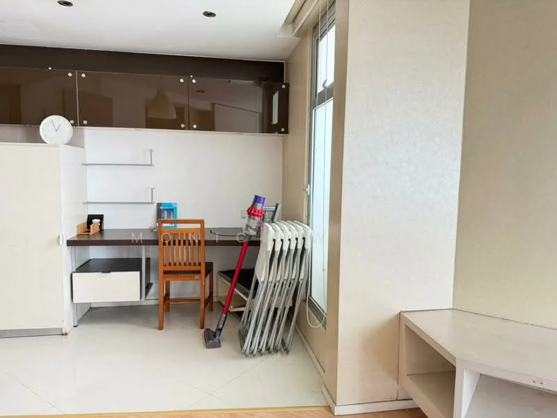 Room features: white painted walls, light-colored flooring, large window with natural light, desk with chair, wooden chair, vacuum cleaner, radiator, shelf unit, clock on wall, minimalist decor; style: modern.