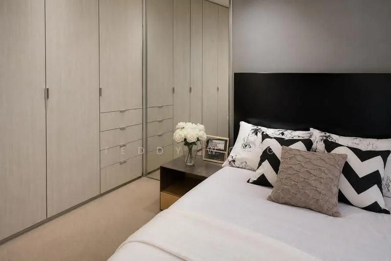 Bedroom features: queen-size bed with white bedding, black upholstered headboard, decorative pillows with geometric patterns, nightstand with a glass top, framed photo on nightstand, built-in wardrobe with sliding doors, light grey cabinetry, carpeted flooring, neutral wall color, table lamp on nightstand, decorative flowers in a vase; style: modern.