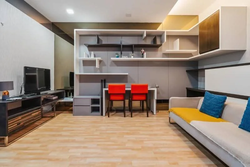 Living features: light wood laminate flooring, grey feature wall, white painted walls, built-in shelving unit with open and closed storage, TV console with storage, wall-mounted TV, 2 red dining chairs, small dining table, beige fabric sofa, blue decorative cushions, yellow accent cushion, table lamp on console; style: modern.