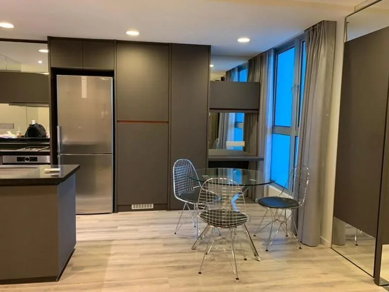 Kitchen features: stainless-steel fridge, built-in oven, 4-burner gas hob, grey cabinetry, black countertop, glass dining table, 4 transparent chairs, light wood flooring, floor-to-ceiling windows, curtains, recessed downlights, mirrored wall; style: modern; view: city.