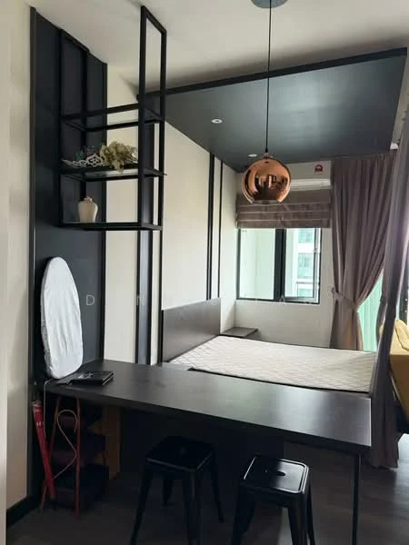 Room features: black metal shelving unit, queen-size bed frame without mattress, black dining table, two black stools, pendant light fixture in copper finish, grey curtains, white walls, dark grey ceiling, window with city view, iron board, decorative plant on shelf; style: modern; view: city.