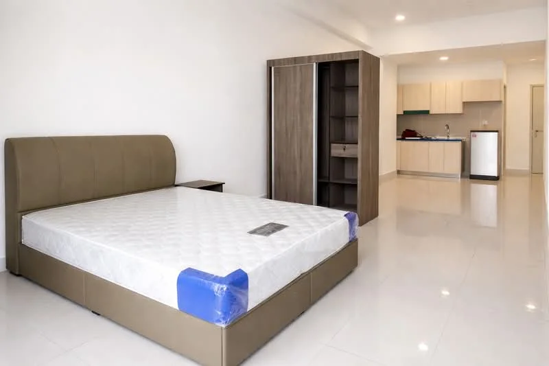 Bedroom features: queen-size bed with mattress, upholstered headboard, white bedding, built-in wardrobe with sliding doors, white painted walls, glossy tile flooring, recessed ceiling lights, nightstand, kitchenette visible in background, stainless-steel fridge visible in background; style: modern.