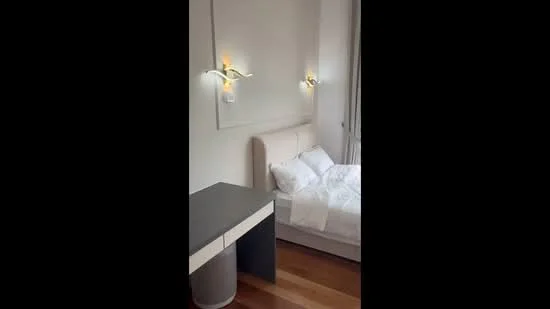 Bedroom features: queen-size bed with white bedding, upholstered headboard, two wall-mounted sconces, wooden flooring, side table with drawer, large window with natural light, decorative wall panel, neutral wall color; style: modern.