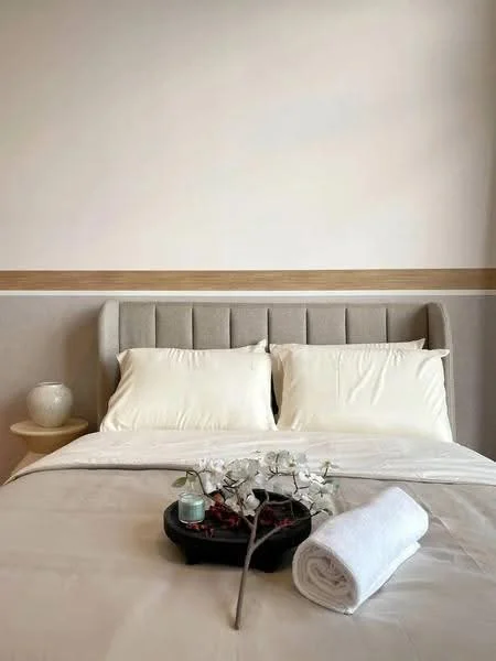 Bedroom features: king-size bed with upholstered headboard, white bedding, decorative tray with flowers, white rolled towel, nightstand with lamp, plain white walls, wood accent strip on wall, soft lighting; style: modern.
