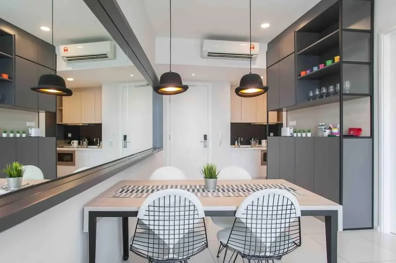 Dining features: white dining table, 4 white wire-frame dining chairs, black pendant lights, grey cabinetry, black countertop, kitchen area visible in background, 2 wall-mounted split AC units, mirror wall, decorative plant on table, open shelving with colored cups, light wood cabinetry, white tiled flooring; style: modern.