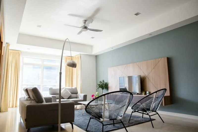 Living features: 3-seater fabric sofa in grey, 2 rattan accent chairs, coffee table, wall-mounted TV, TV console with wood paneling, area rug, floor-to-ceiling windows, sheer curtains, 5-blade ceiling fan, recessed downlights, decorative floor lamp, green potted plant, decorative throw pillows; style: modern; view: city.