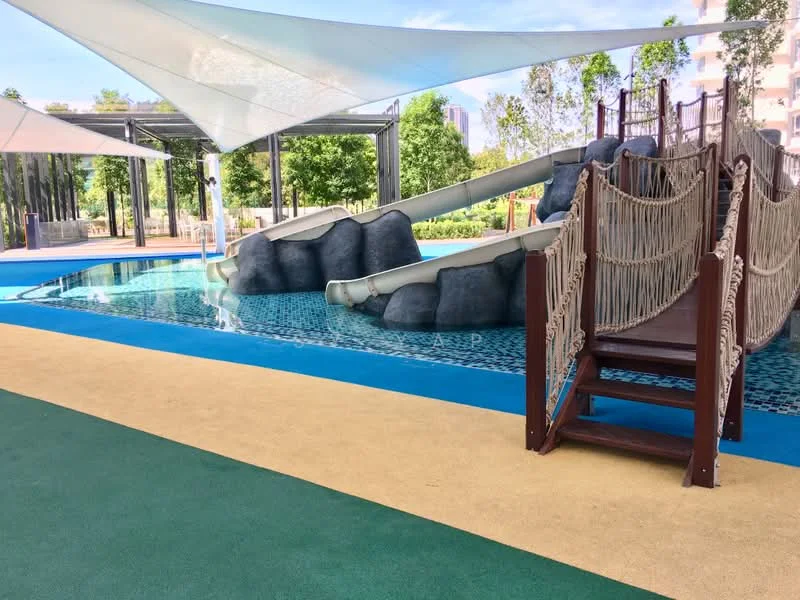 Exterior type: landscape garden; features: swimming pool with water slides, rock formation feature, sandy beach area, shaded seating area with canopies, lush greenery surrounding the pool, wooden bridge leading to slides, blue tiled pool surface, non-slip flooring around the pool; style: tropical-modern.
