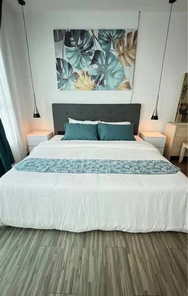 Bedroom features: king-size bed with upholstered headboard, white bedding with decorative throw, two bedside lamps with pendant lighting, framed artwork on wall, wooden flooring, curtains in dark green, dressing table, wall-mounted mirror; style: modern.