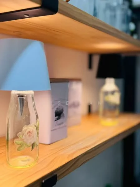 Room features: wooden shelving, glass bottle with floral design, white lamp with shade, decorative box, glass bottle with yellow liquid, ambient lighting; style: modern.