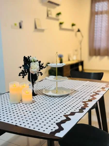 Dining features: black dining table, white tablecloth with black polka dots, decorative cake stand, artificial flower arrangement, three candles, black dining chairs, wall-mounted shelves, curtains on window, white walls, tile flooring; style: modern.