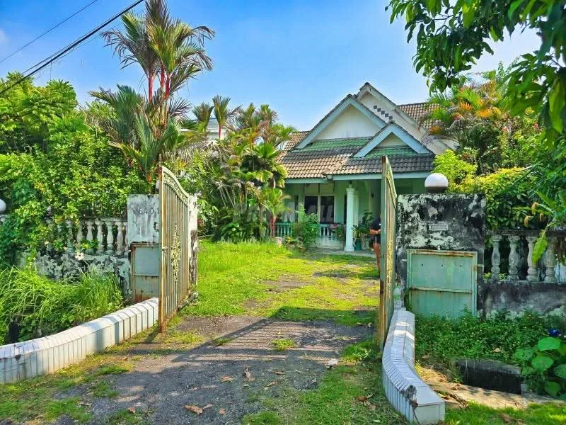 Exterior type: detached bungalow; features: gated entrance, manicured lawn, tropical landscaping, paved walkway, single-storey, sloped roof, porch area, surrounded by greenery, exposed brick facade, white painted fence, decorative stone wall; style: tropical-modern.