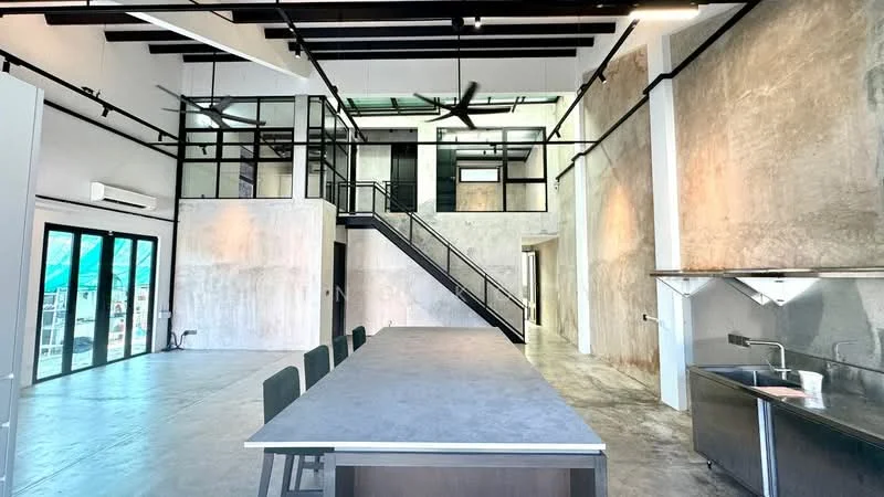 Room features: exposed concrete walls, grey polished concrete flooring, high ceiling with exposed beams, large floor-to-ceiling windows, black metal staircase, open-plan layout, ceiling fan, industrial-style lighting fixtures, kitchen island with grey countertop, stainless-steel sink, stainless-steel countertop, black dining chairs, glass partition, air conditioning unit; style: industrial.