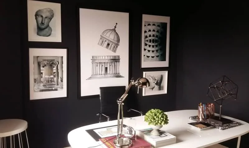 Study features: black painted walls, white desk, metal desk lamp, decorative plant in ceramic pot, framed artwork, geometric sculpture, books and magazines on desk, black office chair; style: modern.