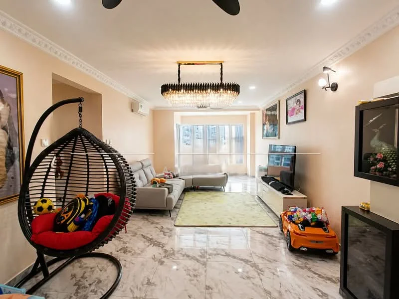 Living features: marble tile flooring, cream painted walls, chandelier, 5-blade ceiling fan, floor-to-ceiling windows, grey fabric sofa, TV console, wall-mounted TV, area rug in green, hanging rattan chair, decorative wall art, toys scattered on the floor, black TV cabinet; style: modern.