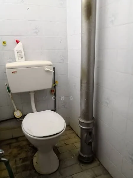 Bathroom features: white toilet, chrome flush handle, white tiled walls, grey tiled flooring, visible plumbing pipe, cleaning product bottle on tank, toilet paper holder, wall-mounted faucet, small drainage pipe.