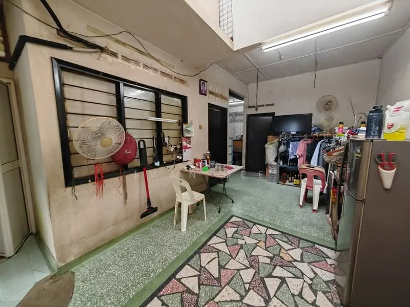 Kitchen features: green terrazzo flooring, white painted walls, exposed ceiling, window with black metal grille, standing fan, refrigerator, kitchen table with patterned surface, plastic chairs, clothes rack, cleaning supplies, various kitchen items on shelves.