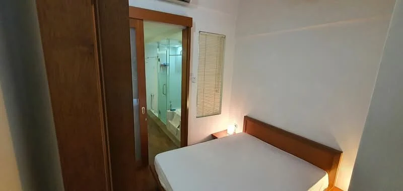 Bedroom features: wooden bed frame, empty mattress, white walls, laminate flooring, built-in wardrobe with sliding doors, window with blinds, door leading to bathroom, ceiling-mounted light fixture; style: modern. Bathroom features: shower area, glass shower enclosure, white tiled walls, tile flooring, sink with vanity, mirror above sink, toilet, light fixture; style: modern.