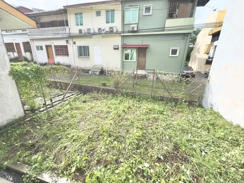 Exterior type: linked terrace house; features: fenced yard, overgrown grass, adjacent houses, multiple air conditioning units visible, green painted facade, wooden door, windows with grills, concrete pathway; style: traditional.