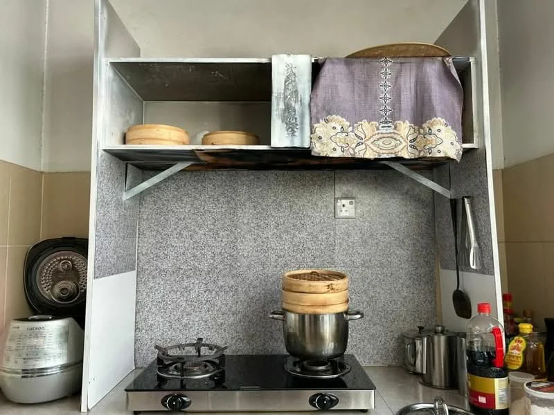 Kitchen features: stainless-steel gas stove with two burners, stainless-steel pot, wooden steamer, grey granite countertop, metal shelving above stove, decorative cloth covering on shelf, various cooking condiments, kettle, rice cooker, wall-mounted electrical outlet; style: modern.